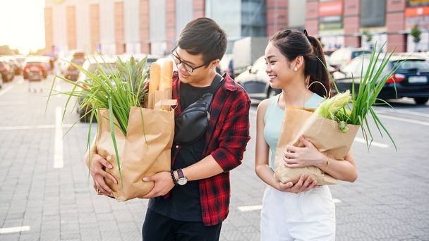 Handsome smiling guy in glasses and cute slender girl go with two big paper bags full of various products purchased in the store - Фото, зображення
