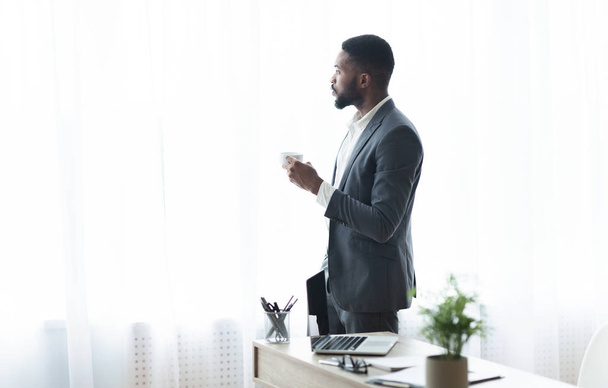 Ευγενικός μαύρος επιχειρηματίας που κοιτάζει έξω από το παράθυρο και πίνει καφέ - Φωτογραφία, εικόνα
