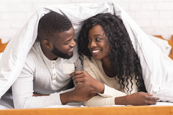 Black couple holding condom, lying on bed under white blanket - Foto, Bild