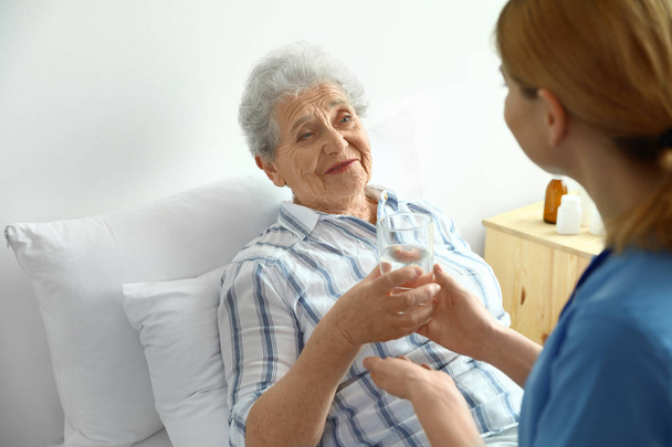 Νοσοκόμα δίνοντας ένα ποτήρι νερό σε ηλικιωμένη γυναίκα σε εσωτερικούς χώρους. Ιατρική βοήθεια - Φωτογραφία, εικόνα