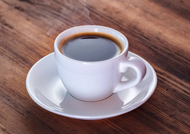 Close up view on smoking coffee in white cup. White coffee cup and saucer on brown table. Texture of wooden desk. Selective soft focus. Blurred background. - Foto, imagen