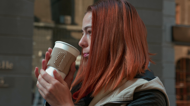 Young attractive red haired woman is drinking hot coffee. Girl with foxy hair drinking tea in paper cup on the street. Autumn cold season. Life in a modern city. Urban lifestyle. Close-up. 4K footage. - Πλάνα, βίντεο