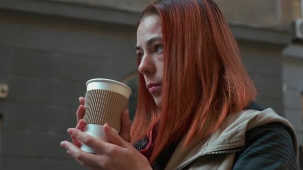 Young attractive red haired woman is drinking hot coffee. Girl with foxy hair drinking tea in paper cup on the street. Autumn cold season. Life in a modern city. Urban lifestyle. Close-up. 4K footage. - Video