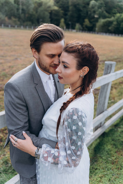 beautiful pregnant woman in white dress with handsome man hugging near wooden fence in field    - Zdjęcie, obraz