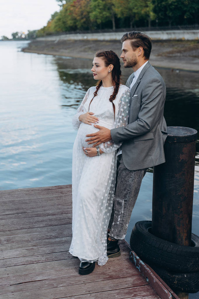 beautiful pregnant woman in white dress with handsome man standing on a wooden pier against lake - Фото, изображение