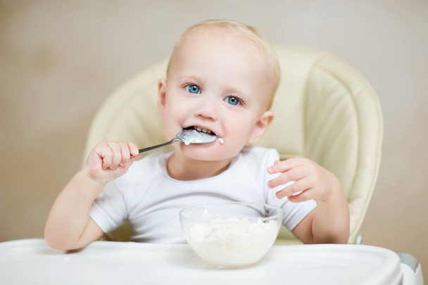 cute baby boy in a chair holds a spoon in his mouth and smiles - Foto, Imagem