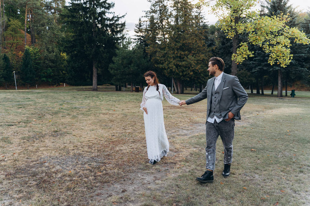 公園で歩く手を握っているハンサムな男と白いドレスで美しい妊娠中の女性 - 写真・画像