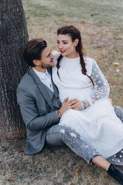 beautiful pregnant woman in white dress with handsome man sitting on the ground near a tree trunk - Fotoğraf, Görsel