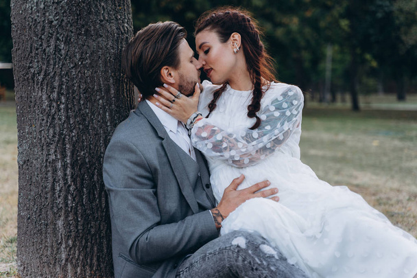 beautiful pregnant woman in white dress with handsome man sitting on the ground near a tree trunk - Valokuva, kuva