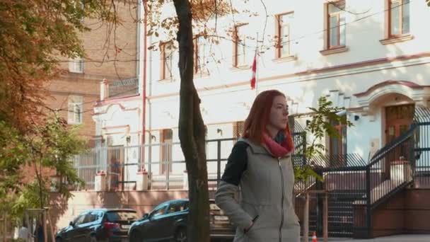 ジャケットを着た若い魅力的な赤い髪の女性が通りを歩いている。セクシーな髪の女の子は晴れた日に屋外で散歩します。秋の暖かい季節。現代都市での生活。好天だ4k映像. - 映像、動画