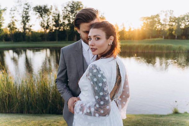 beautiful pregnant woman in white dress with handsome man at green lawn with lake in evening  - Foto, Imagen
