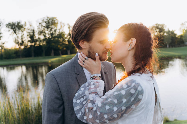 beautiful pregnant woman in white dress with handsome man kissing at green lawn against the setting sun - Foto, Bild