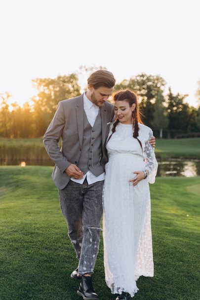 beautiful pregnant woman in white dress with handsome man walking at green lawn against the setting sun - Фото, зображення