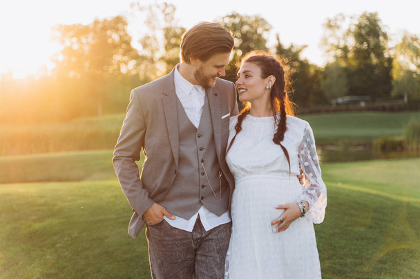 beautiful pregnant woman in white dress with handsome man at green lawn with lake in evening  - Photo, Image