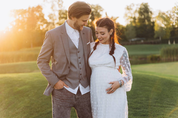 beautiful pregnant woman in white dress with handsome man at green lawn with lake in evening  - Valokuva, kuva