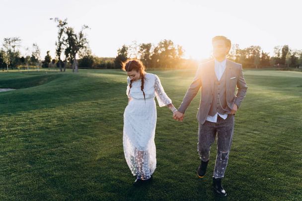 beautiful pregnant woman in white dress with handsome man holding hands and walking at green lawn against the setting sun - Foto, Bild