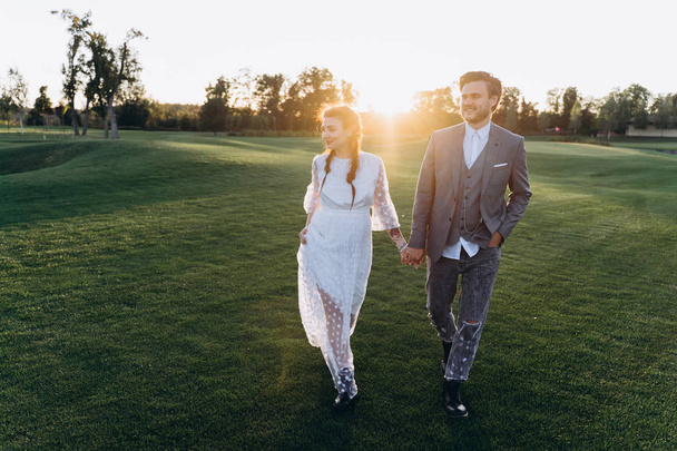 beautiful pregnant woman in white dress with handsome man holding hands and walking at green lawn against the setting sun - Photo, image