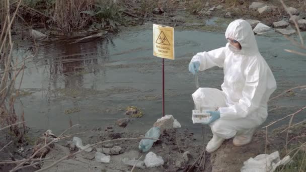 Biyolojik tehlike acil durumu, üniformalı tehlikeli madde işçisi zehirli gölde biyolojik tehlike işareti ile test edilmek üzere test tüplerinde enfekte su örneği alıyor. - Video, Çekim