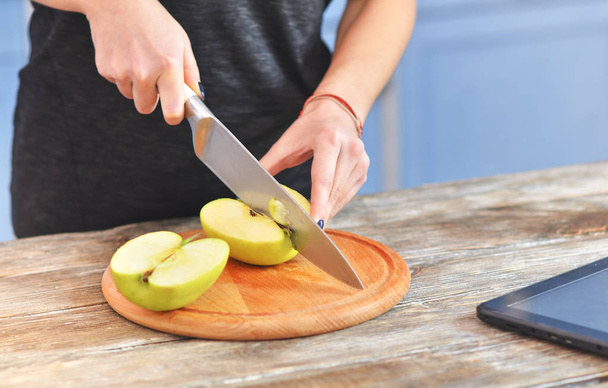 A young woman in a black t-shirt cuts green apple on a light wooden board in a kitchen. - Photo, image