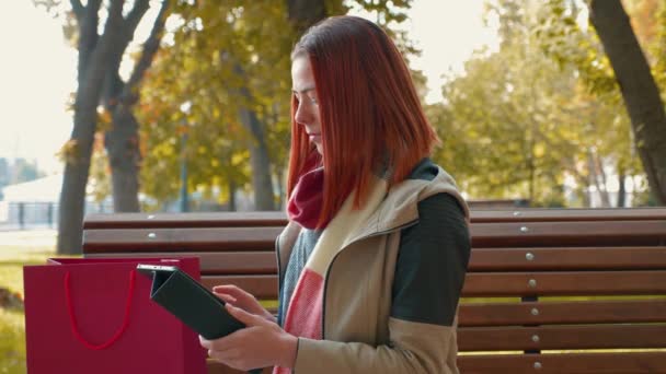 Jovem atraente mulher de cabelos vermelhos em uma jaqueta no parque está comprando online. Menina com cabelo foxy sentado no banco de sol usar tablet tempo e faz um pedido on-line. Venda, conceito da indústria de varejo. 4K
 - Filmagem, Vídeo