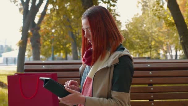 Jonge aantrekkelijke roodharige vrouw in een jasje in het park is online aan het winkelen. Meisje met foxy haar zittend op bank bij zonnig weer gebruik tablet en maakt een online bestelling. Verkoop, retail industrie concept. 4k - Video