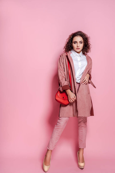 Happy curly brunette girl in coat and bag posing on pink background - Photo, Image