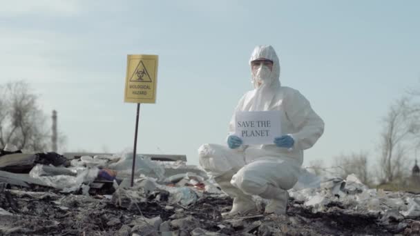 Zanieczyszczenie środowiska, Hazmat człowiek w mundurze pokazuje znak uratować planetę na wysypisku ze wskaźnikiem zagrożenia biologicznego - Materiał filmowy, wideo