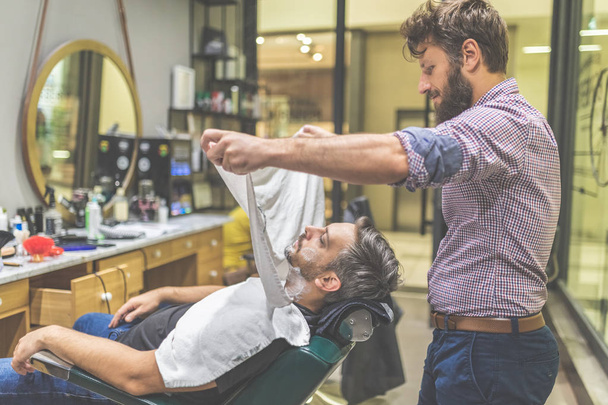 Barber covering client with towel before giving him a shave at barbershop. - Foto, afbeelding