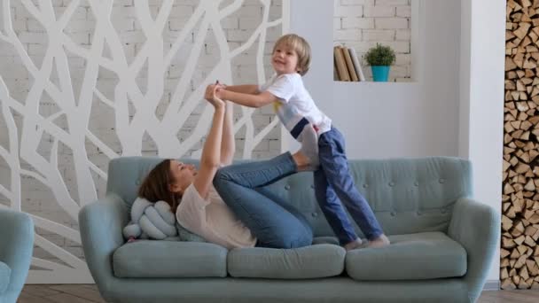 Cheerful mom is playing with her son by lifting him up like a plane. Child having fun with mother. Indoors - Кадри, відео