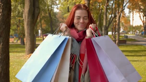 Fiatal vonzó vörös hajú nő kabátban vásárol. Boldog lány róka haj sétál a parkban vásárlások többszínű papírzacskók és örül. Eladó, kiskereskedelmi iparág koncepció. 4k felvétel. - Felvétel, videó