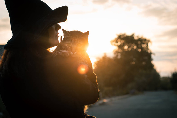 Gato de Halloween en las manos y una bruja con un sombrero negro y un abrigo en una puesta de sol de otoño. Concepto de celebración y mascotas
. - Foto, imagen