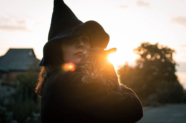 Хэллоуинская кошка в руках и ведьма в черной шляпе и пальто на осеннем закате. Концепция праздников и домашних животных
. - Фото, изображение