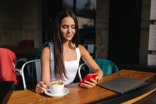 Молодая деловая леди работает на ноутбуке и пьет кофе. Красивая девушка сидит в кафе на улице. Женщина по телефону
 - Фото, изображение