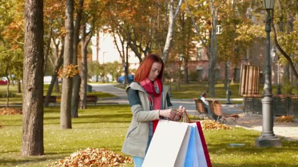 Young attractive red haired woman in a jacket is shopping. Happy girl with foxy hair runs through the park with purchases in multi-colored paper bags and rejoices. Sale, retail industry concept. 4K - Footage, Video