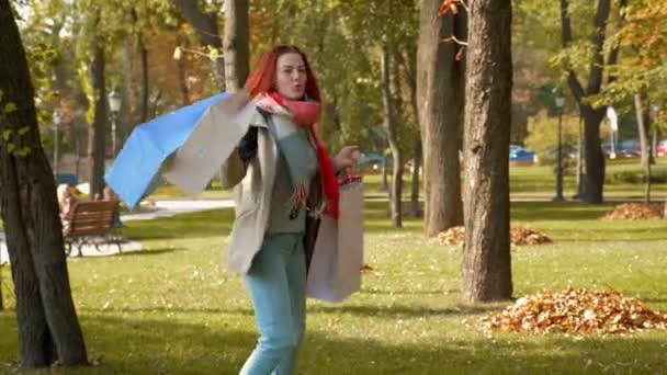Jonge aantrekkelijke roodharige vrouw in jasje is aan het winkelen. Gelukkig meisje met foxy haar wandelingen in het park met aankopen in veelkleurige papieren tassen en vreugde. Verkoop, retail industrie concept. 4k-beelden. - Video