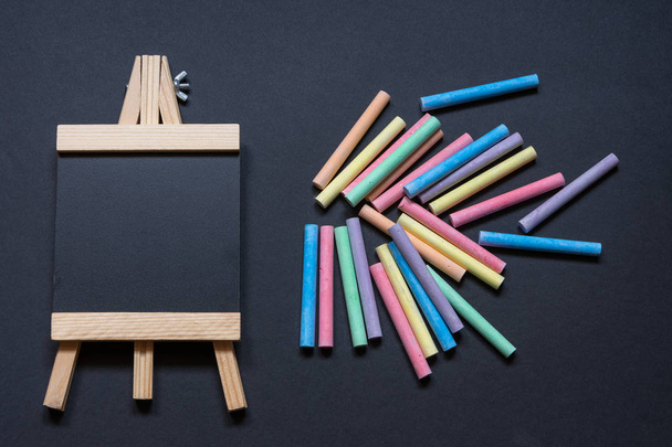 Aerial view of an empty blackboard shaped like an easel on a black background with many messy colored chalks - Photo, Image