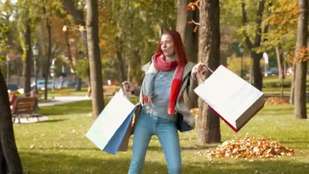 Young attractive red haired woman in jacket is shopping. Happy girl with foxy hair dancing in the park with purchases in multi-colored paper bags and rejoices. Sale, retail industry concept. 4K - Footage, Video