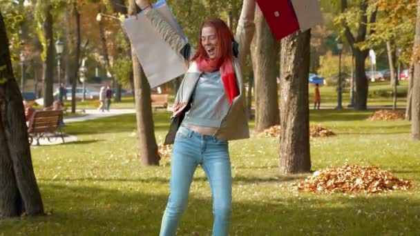 Ceketli, çekici, kızıl saçlı kadın alışveriş yapıyor. Parkta dans eden seksi saçlı mutlu bir kız. Çok renkli kağıt torbalarla alışveriş yapıyor ve seviniyor. Satış, perakende sektörü konsepti. 4k - Video, Çekim