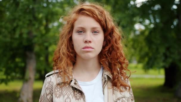 Serious teenager redhead girl looking at camera standing outdoors in park - Video