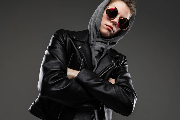 fashionable young woman posing in leather jacket and sunglasses against dark background - Photo, Image