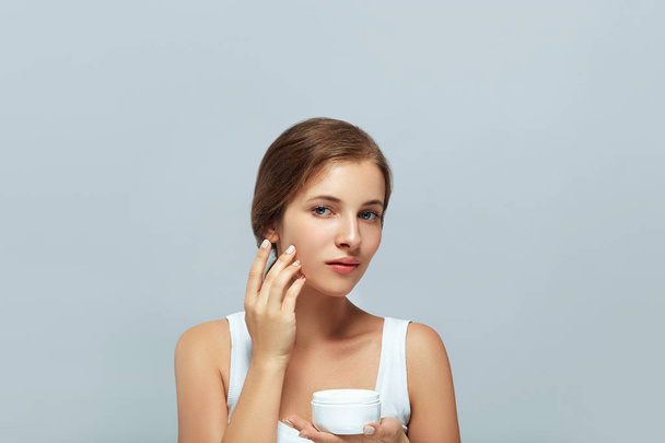 Skincare. Beauty Concept. Young pretty woman holding  cosmetic cream and applying on her face. Soft skin and naked shoulders,model with light nude make-up, portrait of female holding moisturizing cream in her hand and touch own face. Skin Protection  - Foto, Bild