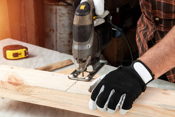 A man carpenter cuts a wooden beam using an electric jigsaw, male hands with an electric jigsaw closeup. Work with wood. - Photo, image