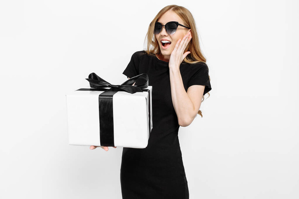 Happy young girl in black dress holding Black Friday gifts. A woman on a white background holds a gift box with a black ribbon - Foto, immagini