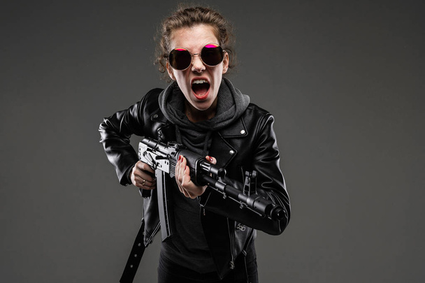 módní mladá žena v kožené bundě a slunečních brýlích pózuje s útočnou puškou proti tmavému pozadí - Fotografie, Obrázek