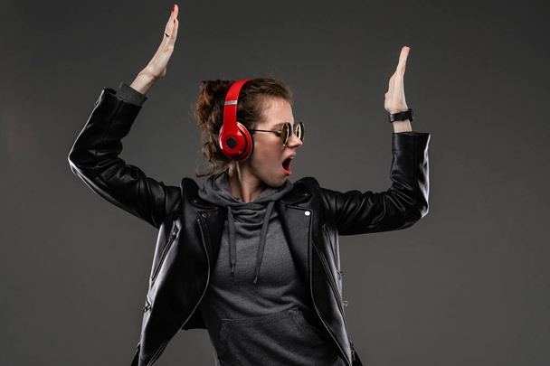 fashionable young woman in red headphones posing in leather jacket and sunglasses against dark background - Fotografie, Obrázek