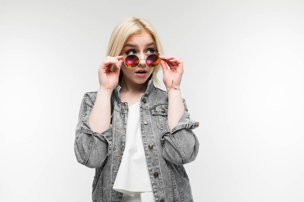 portrait of young charming joyful blonde girl in jeans jacket possing and gesticulating in round sunglasses on background - Photo, image
