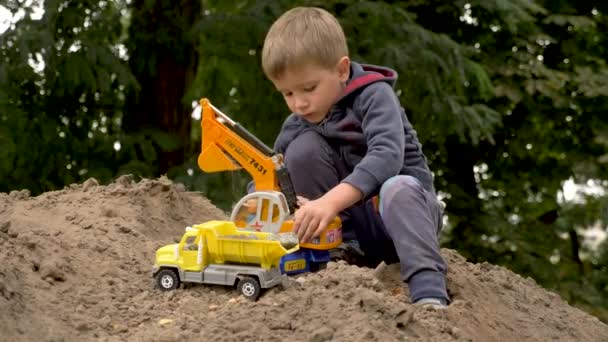 Kind spelen graafmachine en dumper in het park, buiten. Kleine bouwer laadt zand, aarde, gemalen op een vuilniswagen. Bouwconcept. Jongen die met speelgoed speelt voor bouwers. Roll games achtergrond - Video