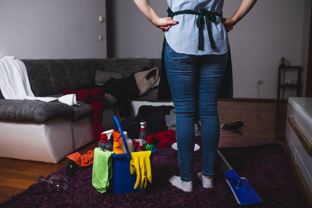 Cansada jovem mulher de pé na sala de estar com produtos de limpeza e equipamentos. Conceito de trabalho doméstico
. - Foto, Imagem