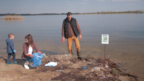 ochrana životního prostředí, chlapec pomáhá rodinným dobrovolníkům čistit znečištěné říční pláže od plastových odpadků a sbírat v pytli na odpadky šetřící ekologii přírody - Záběry, video