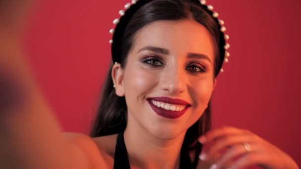 young brunette girl with bright holiday make-up, takes pictures of herself on the phone in the studio. Girl makes selfie on camera. Red background - Záběry, video
